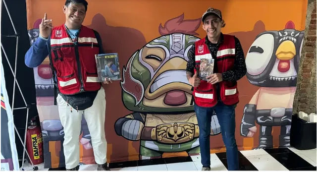 Dos vendedores de la revista Mi Valedor con chaleco rojo sostienen ejemplares frente a un mural de luchadores, en la feria de creadores de El Fucking Hype en Casa Lucerna
